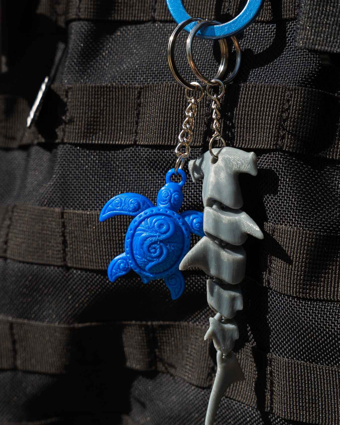 A close-up of a keychain with two charms hanging from metal rings: a bright blue sea turtle charm and a gray shark charm, shown against a black textured fabric background.