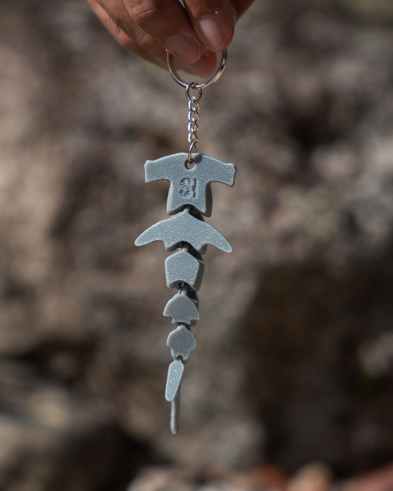 A close-up of a hand holding a Hammerhead Shark Keychain with a stacked, stylized keyring charm against a blurred outdoor rocky background.