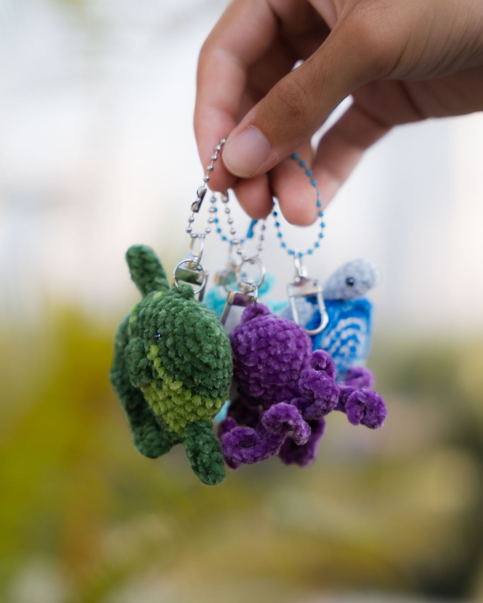 Hand holding a keychain with a green turtle, purple octopus, and blue bird charm against a blurred natural background.